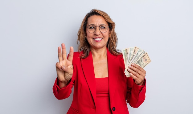 Bella donna di mezza età che sorride e sembra amichevole, mostrando il numero tre. concetto di banconote in dollari