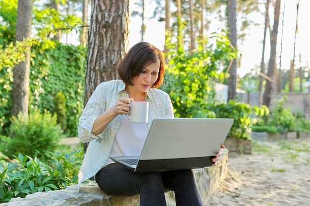 Bella donna di mezza età emotiva che si rilassa in giardino con una tazza di tè e un computer portatile