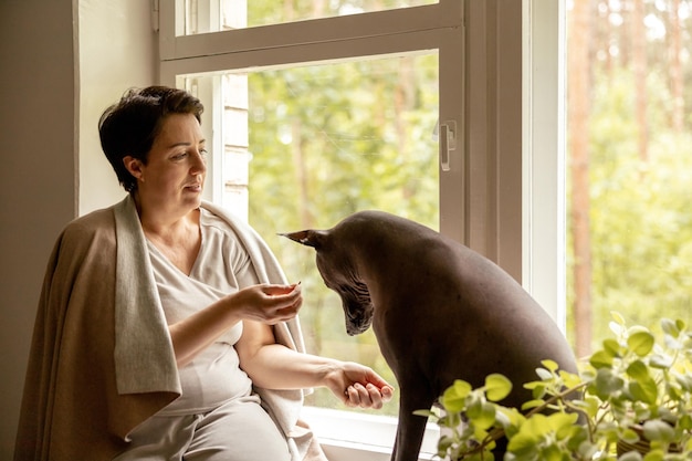 Bella donna di mezza età seduta sul davanzale della finestra con il suo cane donna di 50 anni che trascorre del tempo con il suo cane messicano glabro a casa Xoloitzquintle cane di razza xolo come migliore amico della famiglia