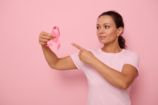 Bella donna di razza mista che indossa una t-shirt rosa che punta a un nastro di raso rosa in mano, sfondo colorato dello spazio della copia. Concetto del 1° ottobre, Giornata internazionale della lotta al cancro al seno