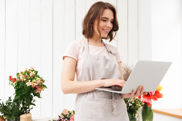 Bella donna di vendita in piedi vicino a mazzi di fiori nel laboratorio di fiori e utilizzando il computer portatile