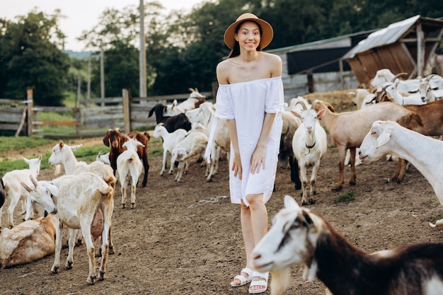 Bella donna e vestito bianco e con un cappello beige tra le capre in una fattoria ecologica