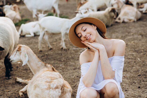 Bella donna e vestito bianco e con un cappello beige tra le capre in una fattoria ecologica