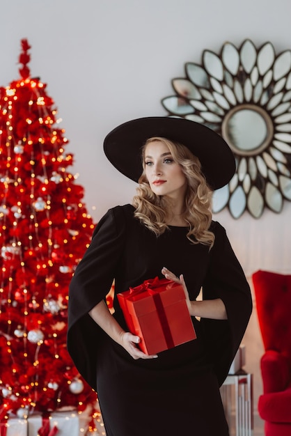 Bella donna elegante in un abito festivo nero e cappello con un regalo di Capodanno sullo sfondo di un albero di Natale rosso Soft focus selettivo