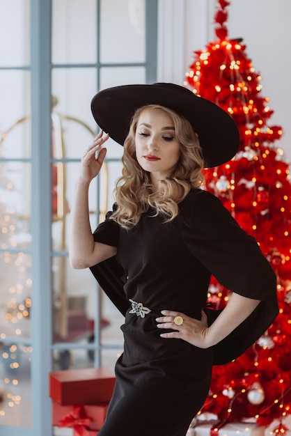 Bella donna elegante in un abito festivo nero e cappello sullo sfondo di un albero di Natale rosso Soft focus selettivo