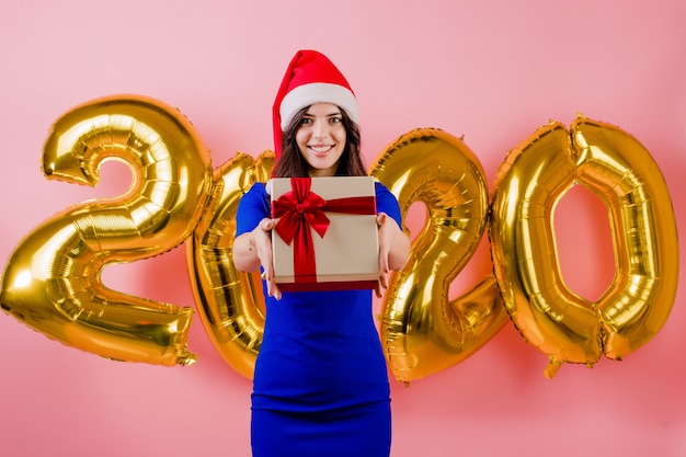 Bella donna emozionante che indossa il contenitore di regalo della tenuta del cappello di Santa davanti ai palloni del nuovo anno 2020 isolati sopra il rosa