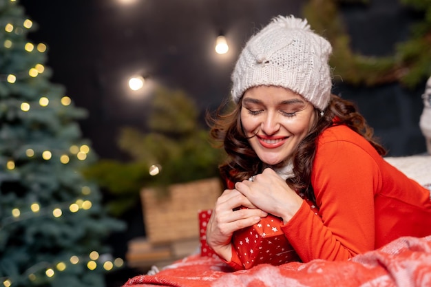 Bella donna felice che tiene il regalo di Natale con gli occhi chiusi Signora in abiti rossi e berretto bianco che tiene presente al suo cuore Bella ragazza sullo sfondo della decorazione di Natale