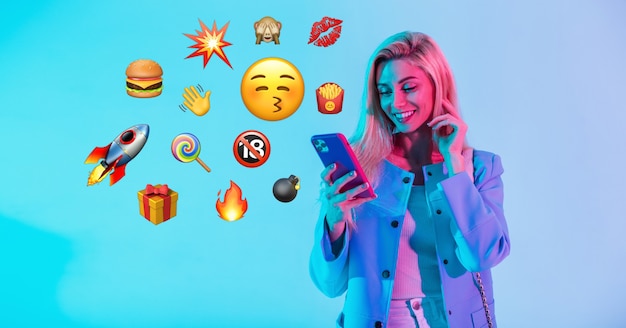 Bella donna felice che tiene smartphone con emoji piatto su sfondo al neon. Social Media Emoji Comunicazione Concept