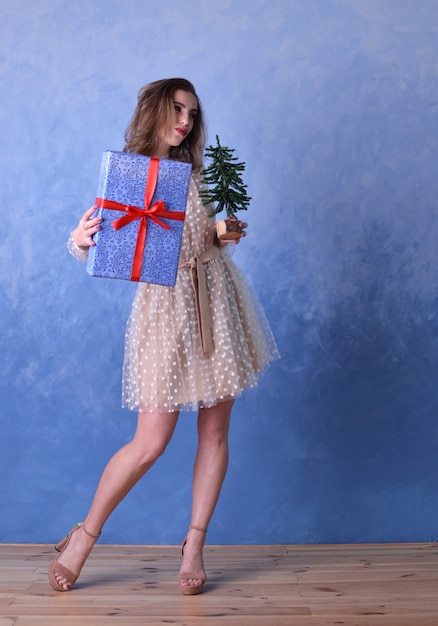 Bella donna felice che tiene un piccolo albero di Natale e un regalo blu con un nastro rosso nelle sue mani