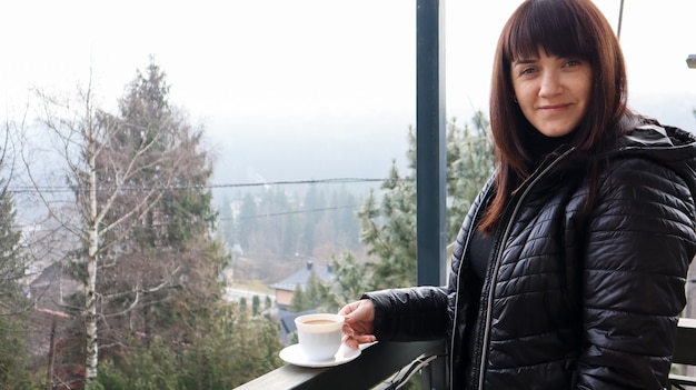 Bella donna felice con una tazza di caffè o tè bianca che si gode la splendida vista del paesaggio di montagna nella nebbia mattutina dal balcone della sua camera d'albergo. Concetto di viaggio autunnale e invernale.