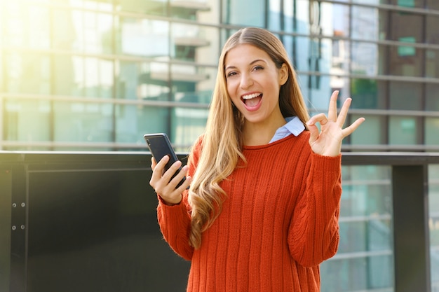 Bella donna felice di autunno che tiene telefono astuto che fa segno giusto alla macchina fotografica