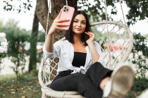 Bella donna felice in abiti di moda con una camicia bianca si siede su un'altalena a maglia e scatta una foto selfie su un telefono rosa