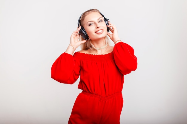 Bella donna felice in vestito rosso alla moda in cuffie che posano sul grigio. La ragazza ascolta la sua musica preferita