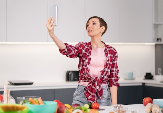 Bella donna felice prendendo selfie utilizzando il suo smartphone in cucina durante la cottura di insalata fresca che indossa una camicia a quadri