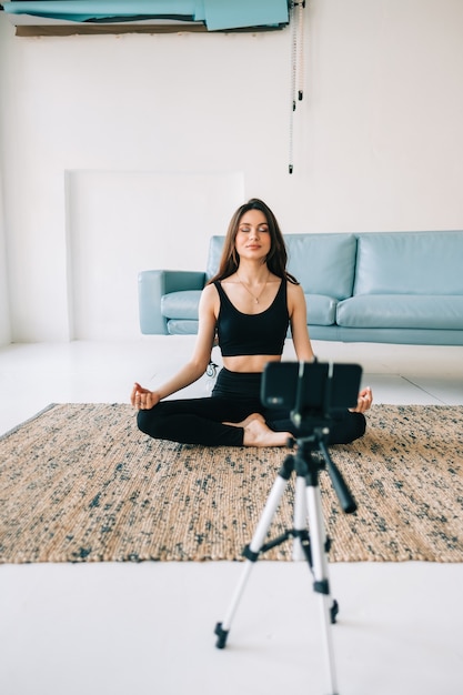 Bella donna fitness meditare nella fotocamera frontale su treppiede, facendo yoga a casa