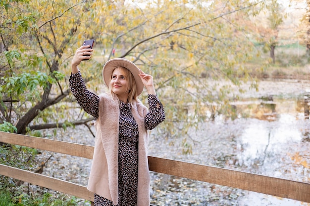 Bella donna in abiti alla moda e cappello prende selfie sul telefono in natura nel parco autunnale in autunno.