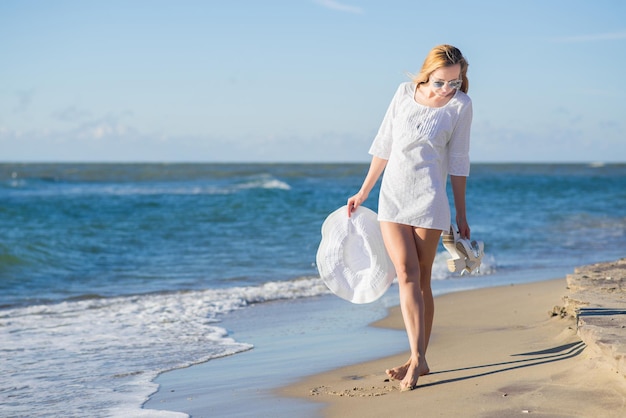 Bella donna in abito bianco che cammina sulla sabbia attraverso la spiaggia in una giornata di sole