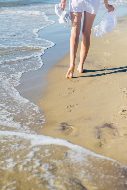 Bella donna in abito bianco che cammina sulla sabbia attraverso la spiaggia in una giornata di sole