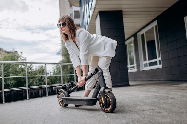 Bella donna in abito bianco che piega il suo scooter elettrico dopo il giro mentre si trova sullo sfondo di un edificio moderno