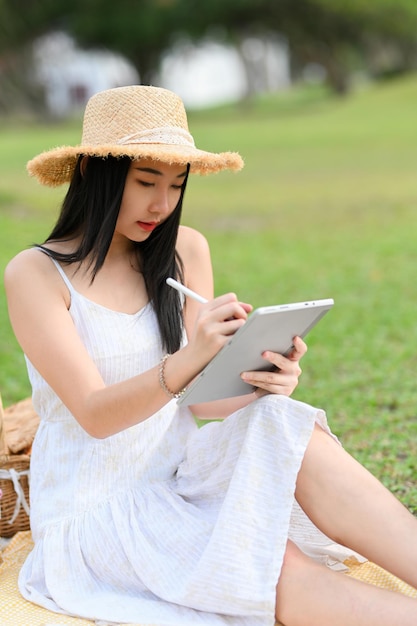 Bella donna in abito bianco e cappello di paglia che ha una giornata di picnic in giardino rilassata utilizzando il tablet