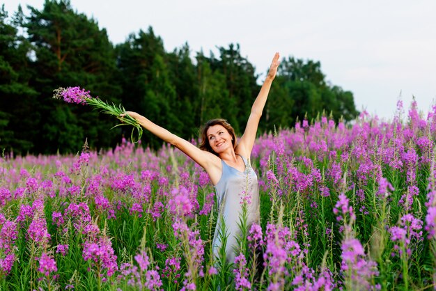 Bella donna in abito grigio esulta con le mani in alto e il mazzo di fiori sul prato di fireweed