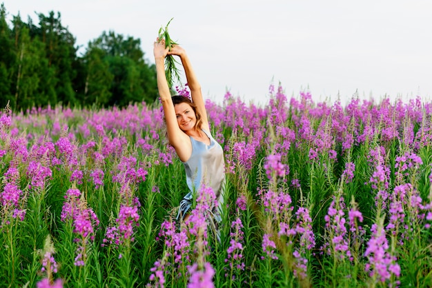 Bella donna in abito grigio esulta con le mani in alto e il mazzo di fiori sul prato di fireweed