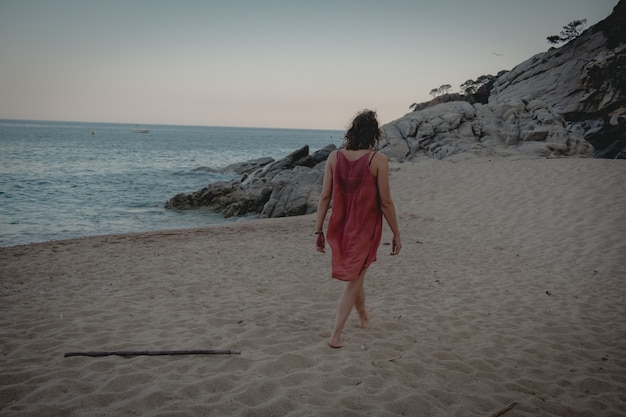 Bella donna in abito lungo arancione sta camminando sulla spiaggia al tramonto