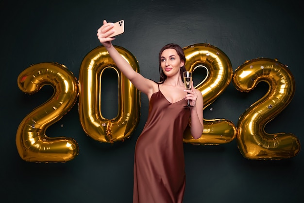Bella donna in abito moderno di lusso che fa selfie con champagne davanti a palloncini dorati isola...