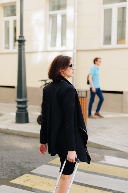 Bella donna in abito nero su uno sfondo di un paesaggio urbano con una valigia