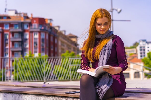 Bella donna in abito viola e sciarpa che legge un libro in un parco della città, immagina e si diverte a leggere