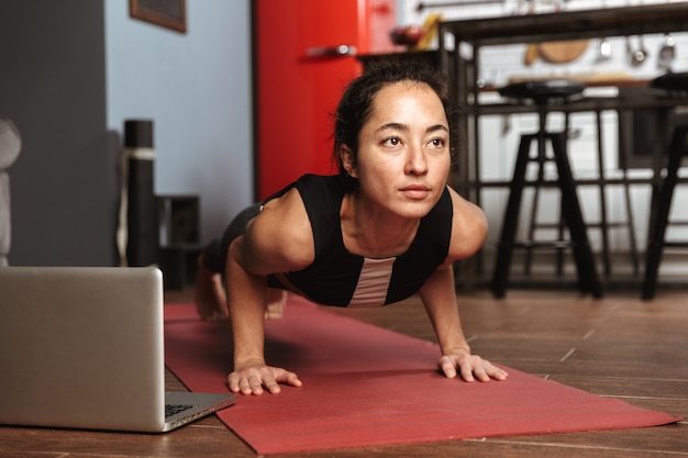 Bella donna in buona salute facendo esercizi di yoga mentre è seduto su un tappetino fitness a casa, utilizzando il computer portatile, stretching