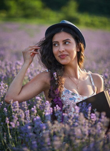 Bella donna in cappello che si siede nel campo di lavanda viola e che tiene un libro.