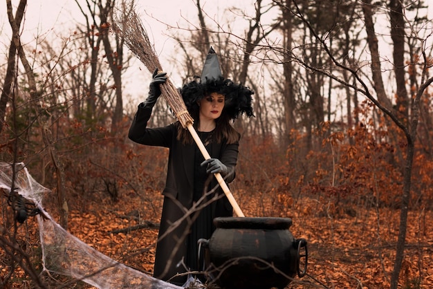 Bella donna in cappello da strega e costume che tiene scopa vicino al calderone nella foresta autunnale Concetto di Halloween Fuoco selettivo