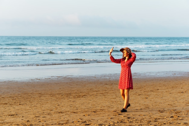 Bella donna in cappello e vestito rosso che prende un selfie o una foto in primavera sulla spiaggia in Spagna