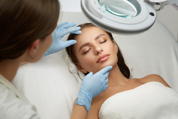 Bella donna in cosmetologia clinica sdraiata con gli occhi chiusi e ottenere test di trattamento viso professionale dal cosmetologo