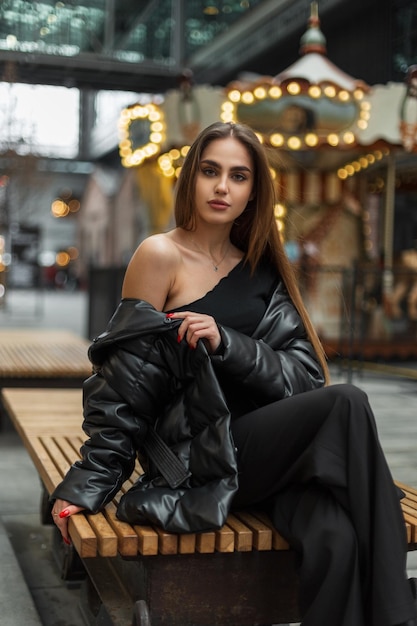 Bella donna in eleganti abiti invernali con piumino si siede sulla strada vicino a una giostra vintage Bellezza e moda in stile femminile urbano