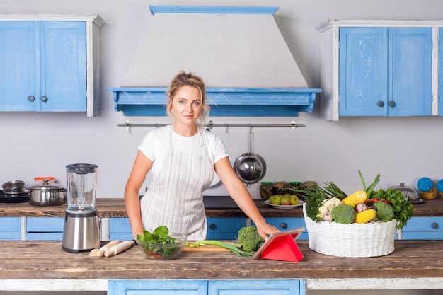 Bella donna in grembiule alla ricerca di ricetta alimentare in internet utilizzando tablet e guardando la fotocamera che cucina insalata in cucina moderna cesto di verdure fresche sul tavolo alimentazione sana dieta vegetariana