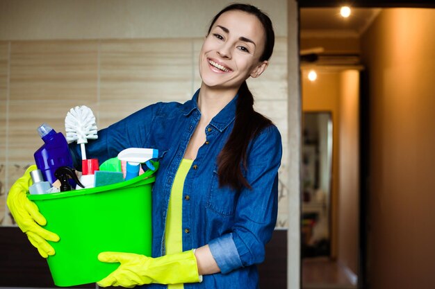 Bella donna in guanti protettivi tiene in mano un secchio con cose per la pulizia guardando la fotocamera e sorridendo prima dell'inizio della pulizia della casa