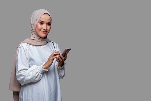 Bella donna in hijab è in possesso di un cellulare sorridendo e guardando la telecamera isolata su sfondo chiaro