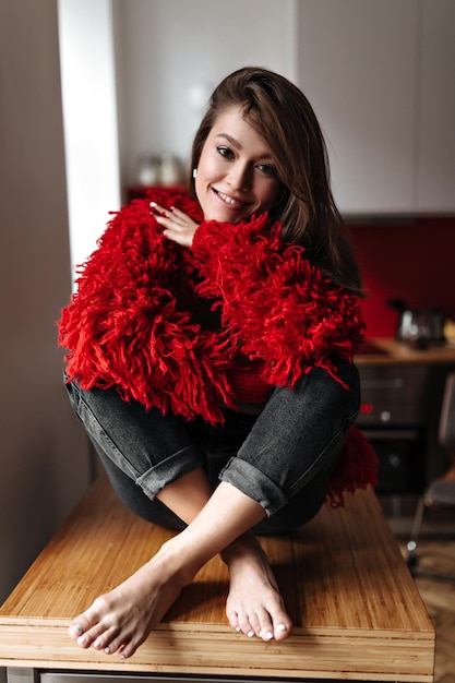 Bella donna in jeans neri e maglione rosso si siede sul tavolo della cucina e sorride dolcemente