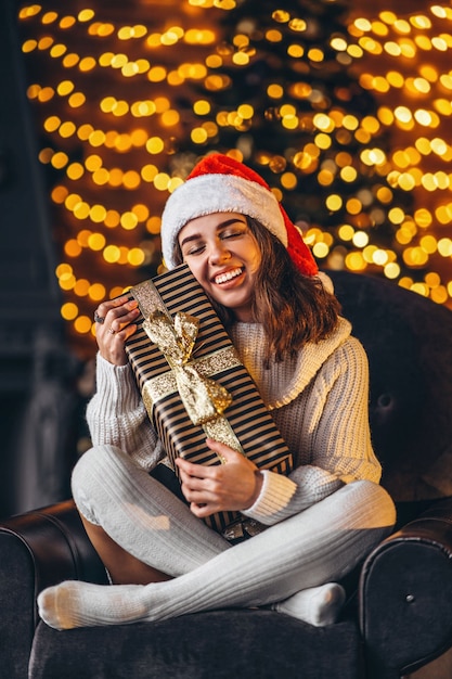 Bella donna in maglione caldo, calzini e cappello di Natale, seduta sulla sedia a casa con confezione regalo