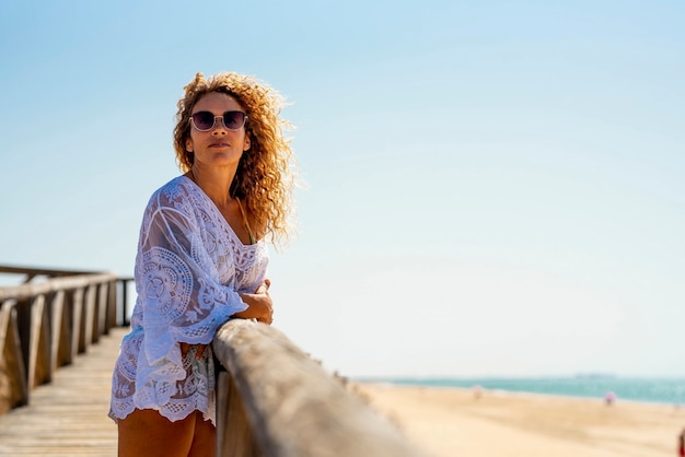 Bella donna in occhiali da sole e abito estivo bianco in piedi sul molo di legno o sul molo sulla spiaggia sabbiosa con vista sul mare contro il cielo blu chiaro. Donna in vacanza in spiaggia