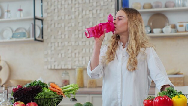 Bella donna in piedi in cucina e beve acqua da una bottiglia sportiva rosa