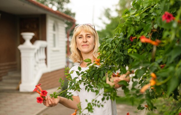 Bella donna in piedi nel cortile vicino al cespuglio con fiori e in posa per la fotocamera Donna bionda adulta che riposa in una casa di campagna in piedi vicino a un bellissimo cespuglio e guardando la telecamera