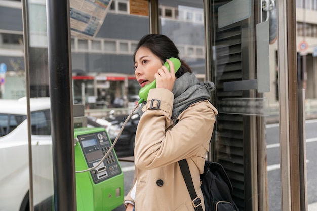 bella donna in piedi nella cabina del telefono e usando il telefono pubblico perché ha dimenticato il suo cellulare a casa