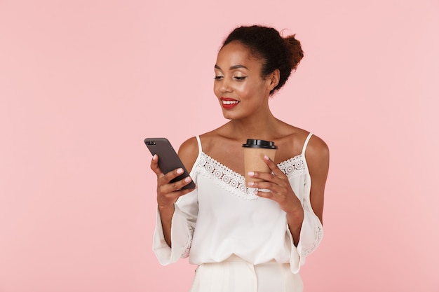 Bella donna in posa isolato sopra la parete rosa bere caffè utilizzando il telefono cellulare e in chat