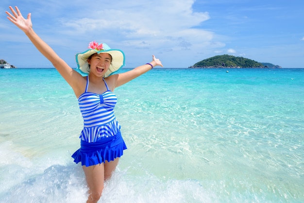 Bella donna in un cappello blu a strisce di costume da bagno in piedi con le braccia tese alla vita spensierata in mare e cielo in estate sulla spiaggia dell'isola di Miang, Parco Nazionale Mu Ko Similan, Phang Nga, Thailandia