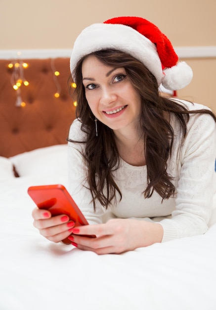 Bella donna in un maglione bianco e cappello di babbo natale in posa con un telefono in mano. Donna che guarda la telecamera e sorride