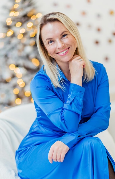 Bella donna in un vestito blu si siede su un letto bianco sullo sfondo delle luci di Natale Ragazza sorridente e guardando la telecamera