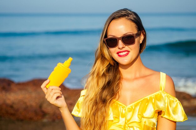 Bella donna in un vestito di seta giallo spruzza uno spray solare sulla spiaggia. concetto di spf pelle perfetta labbra rosse trucco luminoso tropici estate sulla spiaggia